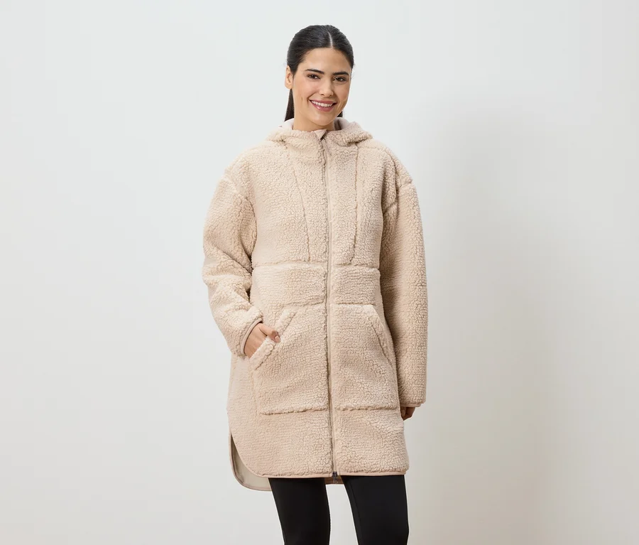 Une femme souriante portant un long manteau en fausse fourrure beige à capuche et des leggings noirs.