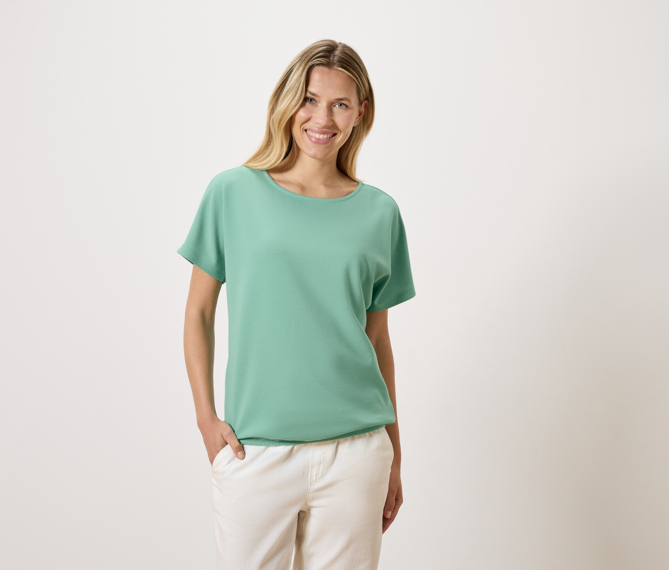 Une femme blonde portant un t-shirt vert et un pantalon blanc sourit.
