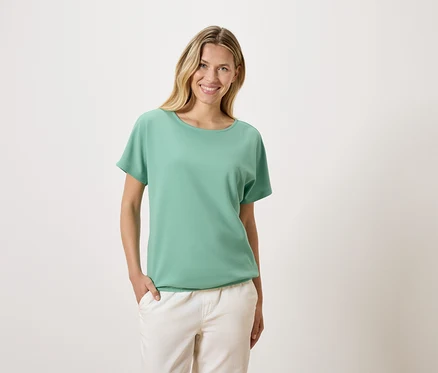 Une femme blonde portant un t-shirt vert et un pantalon blanc sourit.