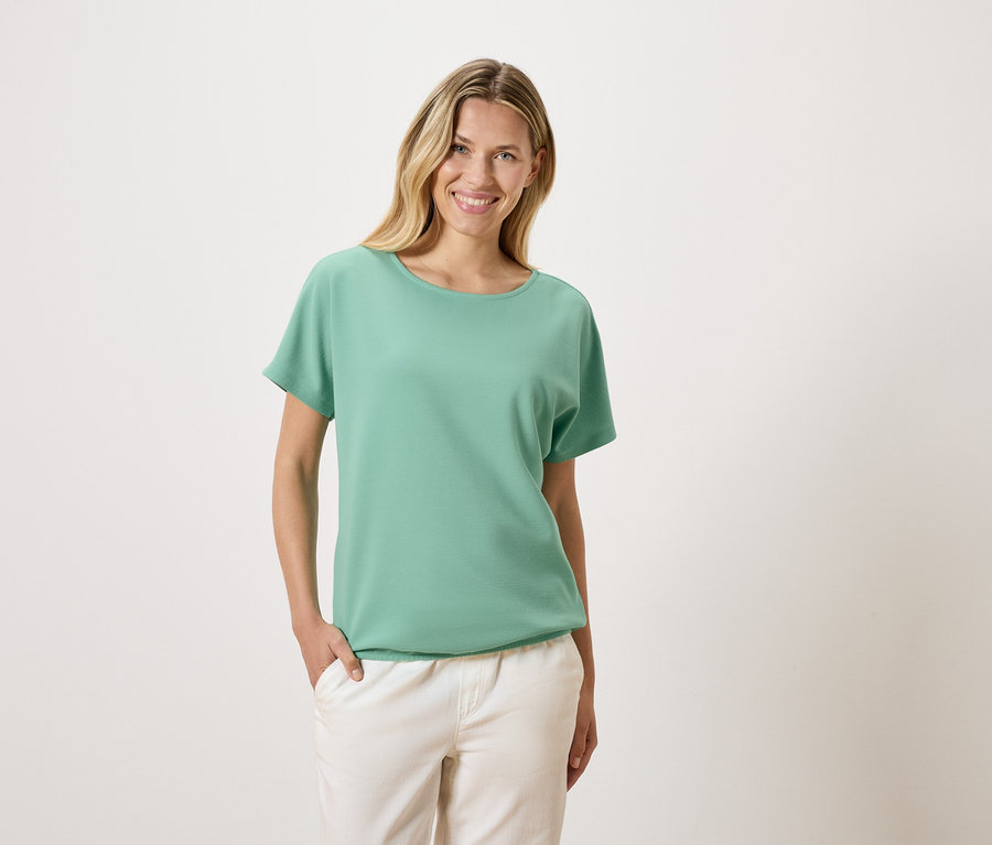 Une femme blonde portant un t-shirt vert et un pantalon blanc sourit.