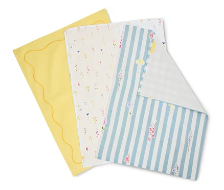 Trois linges sont superposés : un jaune avec un bord ondulé, un crème avec des motifs floraux et un bleu et blanc rayé avec des fleurs.