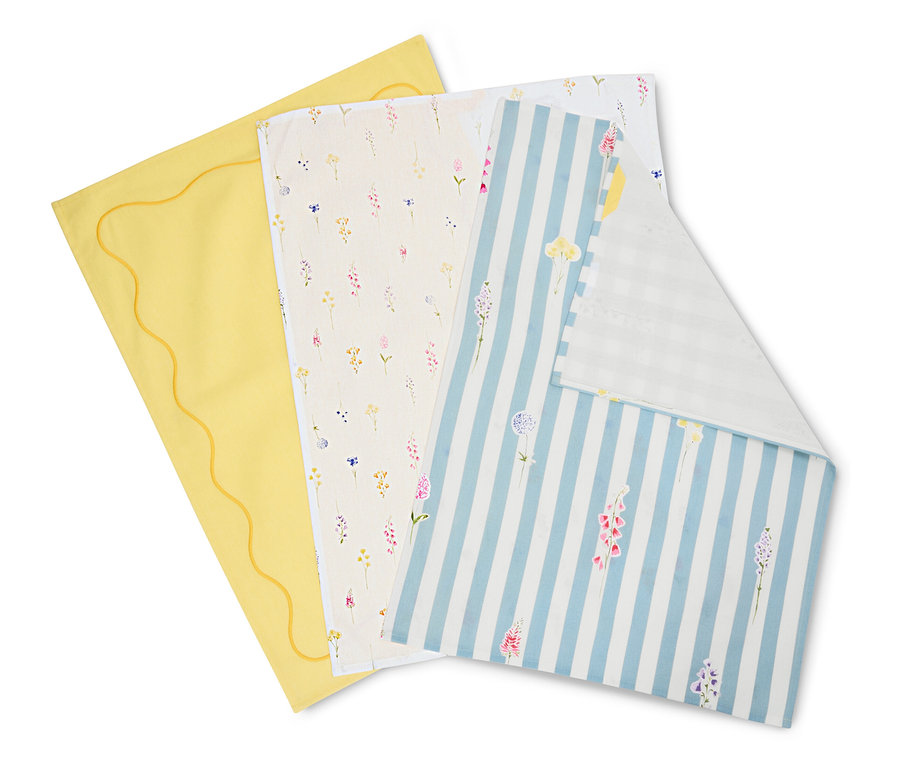 Trois linges sont superposés : un jaune avec un bord ondulé, un crème avec des motifs floraux et un bleu et blanc rayé avec des fleurs.