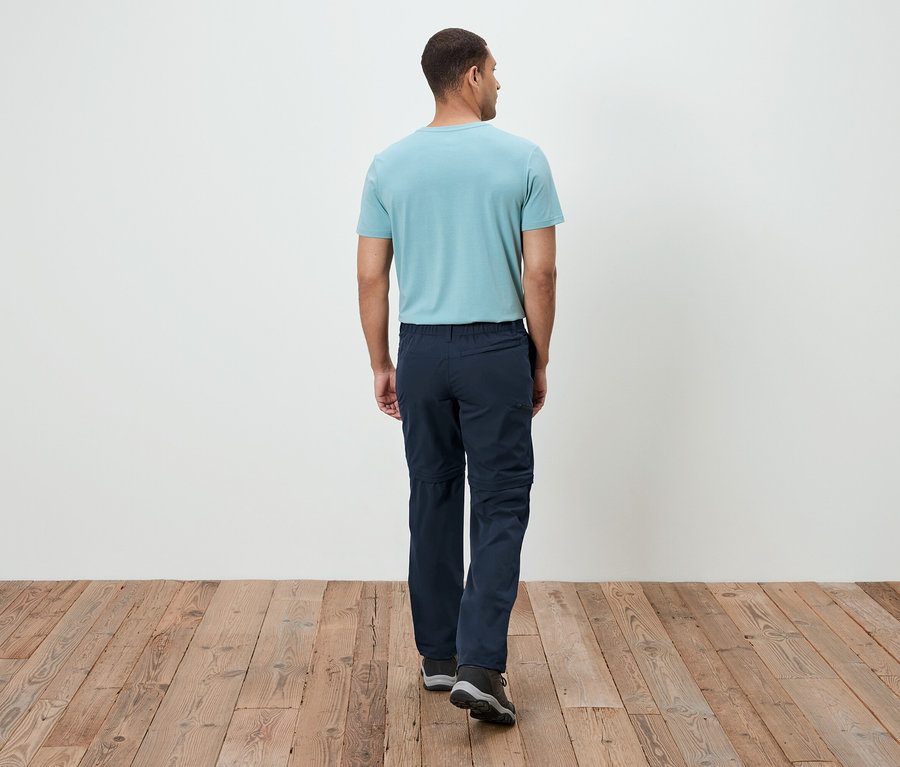 Homme vu de dos, portant un t-shirt turquoise et un pantalon bleu marine, marchant sur un plancher en bois.