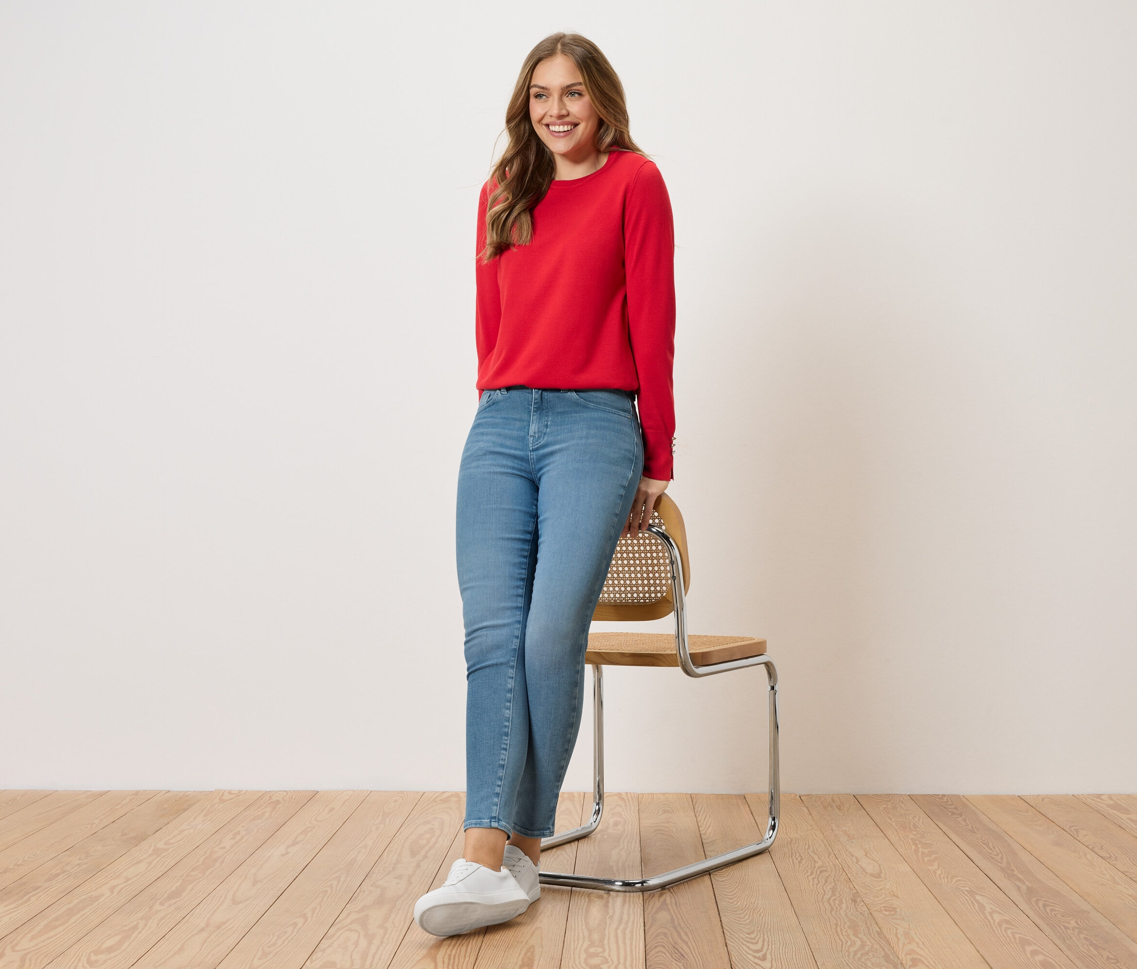 Une femme est appuyée sur une chaise. Elle porte un pull rouge, un jean bleu et des baskets blanches.