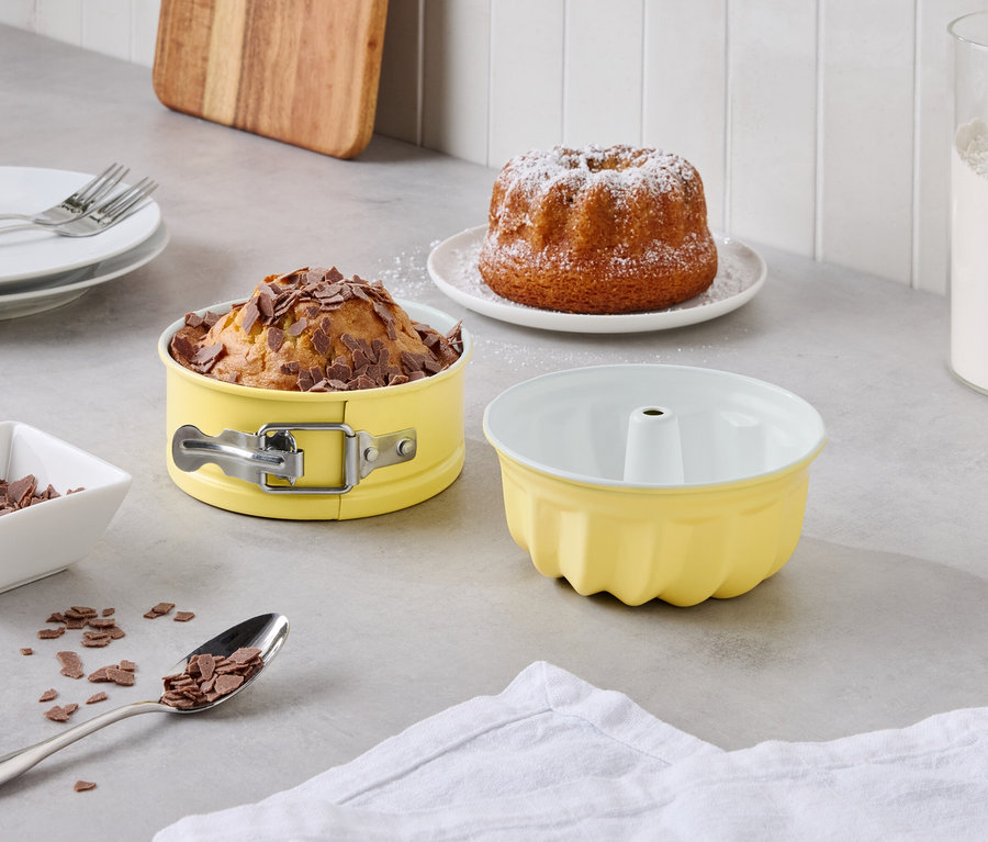 Une table dressée avec un kouglof saupoudré de sucre glace, un gâteau aux pépites de chocolat dans un moule à manqué jaune, un moule à kouglof jaune vide et un bocal de farine.