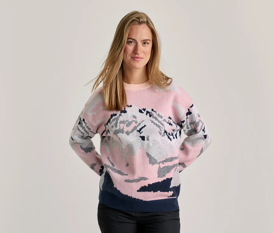 Femme souriante portant un pull rose, gris et bleu foncé avec un motif de montagne, un pantalon noir, mains derrière le dos.