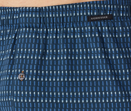 Gros plan sur un tissu bleu avec un motif de rectangles bleus clairs et foncés, une ceinture élastique en haut, une étiquette noire avec « SCHIESSER » et un bouton avec « SCHIESSER EST. 1875 ».