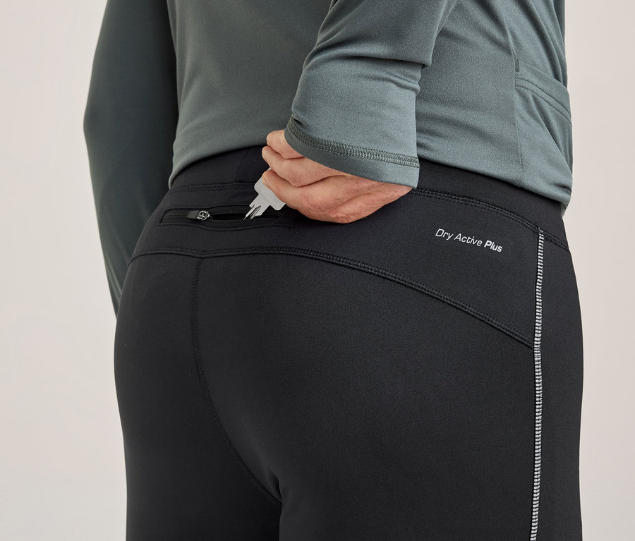 Une personne de dos met des clés dans la poche arrière zippée d'un pantalon de sport noir. Le pantalon porte l'inscription « Dry Active Plus » et une bande réfléchissante.