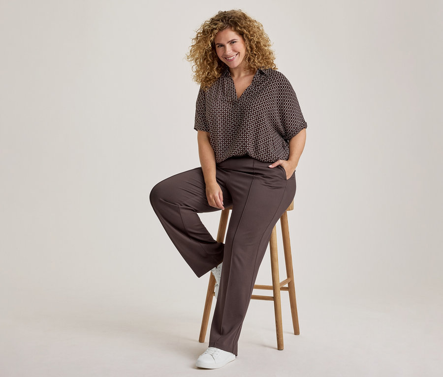 Femme souriante aux cheveux bouclés, vêtue d'une chemise à manches courtes à motifs sombres et d'un pantalon marron foncé, assise sur un tabouret en bois, une main dans sa poche et des baskets blanches aux pieds.