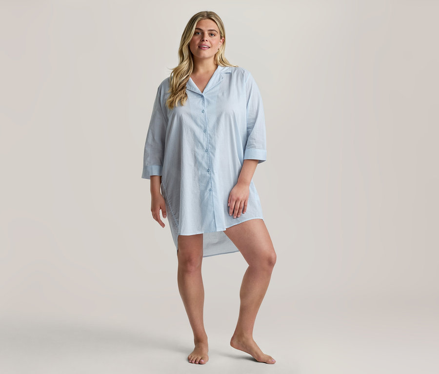 Femme blonde souriante, debout pieds nus, portant une chemise de nuit Web-Nachthemd bleu clair à col, manches trois-quarts et ourlet asymétrique.