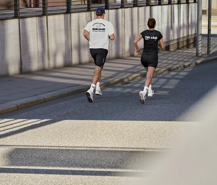 Un homme en t-shirt de sport blanc avec inscription « RUN CLUB », short de sport noir et chaussettes de tennis Unisex Coffee Crew, et une femme en t-shirt de sport noir avec inscription « RUN CLUB » et leggings de sport courts ActivePower courent sur une route.