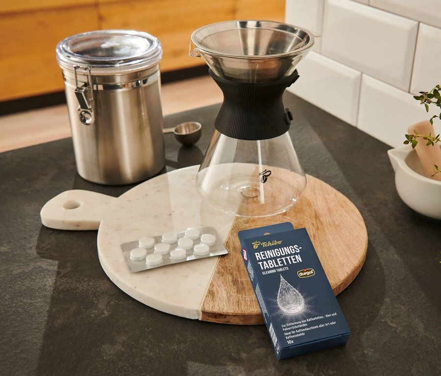 Sur une planche à découper se trouvent des pastilles de nettoyage durgol® pour les huiles de café, un récipient, un filtre à café et une cuillère à mesurer.