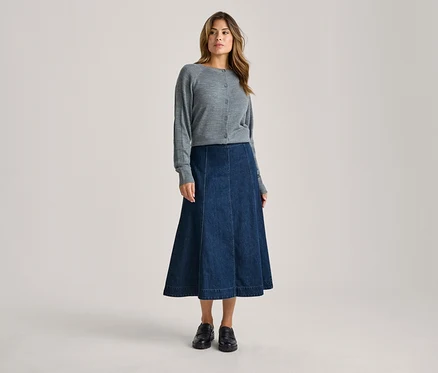 Une femme debout de face, portant un cardigan gris boutonné, une jupe midi évasée en jean bleu foncé et des mocassins noirs.