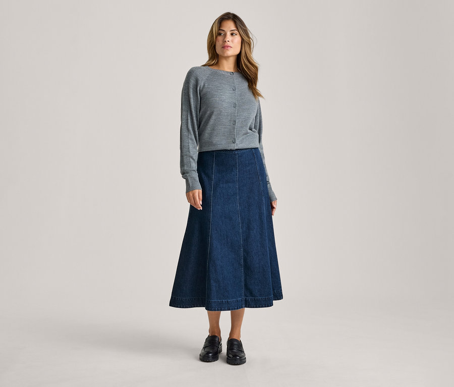 Une femme debout de face, portant un cardigan gris boutonné, une jupe midi évasée en jean bleu foncé et des mocassins noirs.