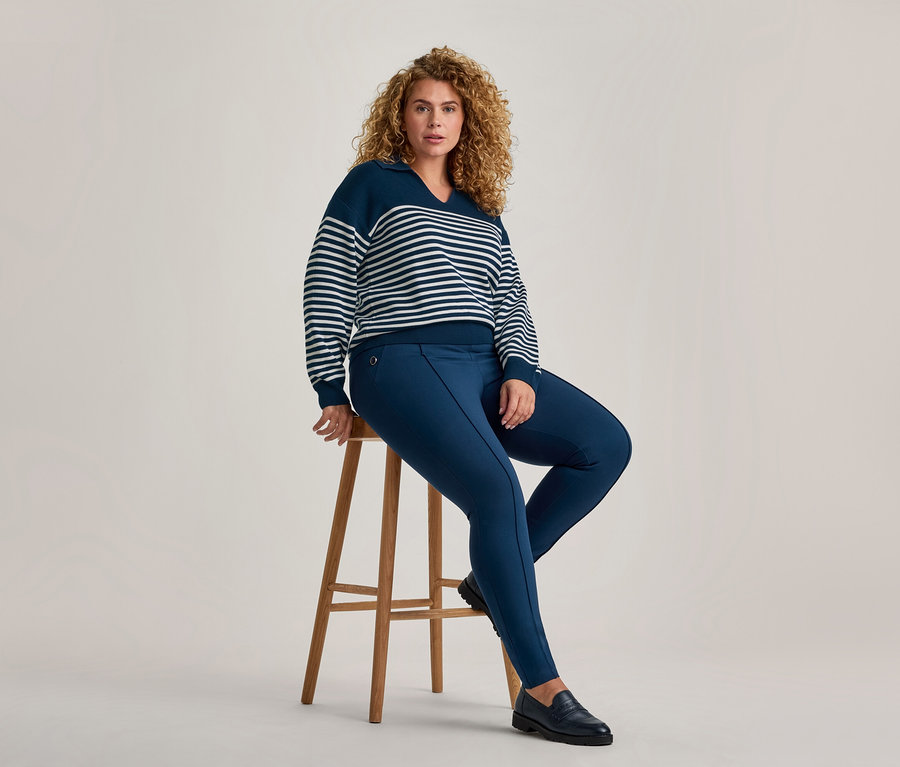 Une femme aux cheveux bruns bouclés est assise sur un tabouret en bois, regardant devant elle. Elle porte un pull rayé bleu marine et blanc à col en V, un pantalon slim bleu marine et des mocassins noirs.