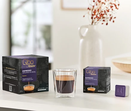 Un verre de Qbo Espresso CAFEZINHO DO BRASIL est placé entre une grande boîte de 27 capsules et une petite boîte de 8 capsules, avec une seule capsule violette à côté de la petite boîte, le tout sur une surface blanche.
