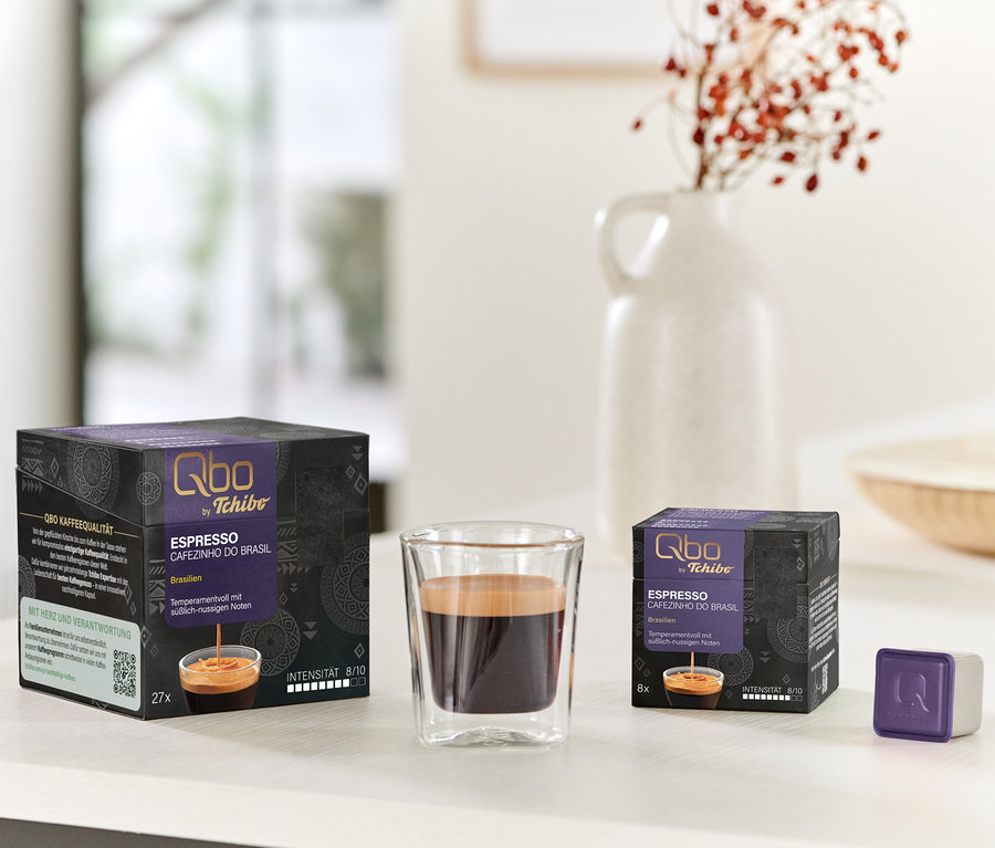 Deux boîtes noires et violettes de capsules Qbo Espresso CAFEZINHO DO BRASIL (27 et 8 unités), un verre d'espresso et une capsule violette sont posés sur une table blanche. Un vase blanc avec des baies rouges est en arrière-plan.