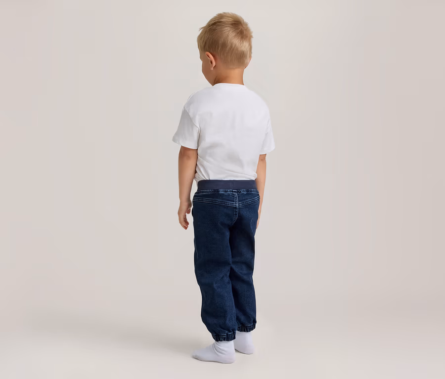 Jeune garçon aux cheveux clairs, debout de dos, portant un t-shirt blanc, un pantalon de jogging bleu foncé et des chaussettes blanches.
