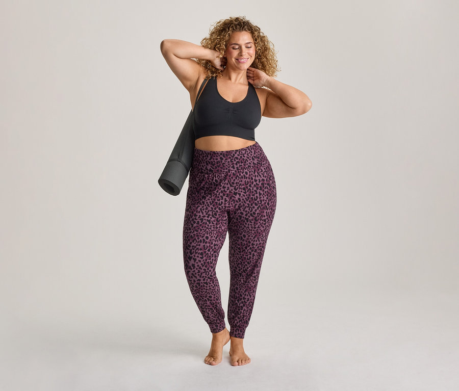 Femme souriante aux cheveux bouclés, vêtue d'un soutien-gorge de sport noir et d'un pantalon de jogging violet à imprimé léopard, tenant un tapis de yoga roulé sur son épaule.