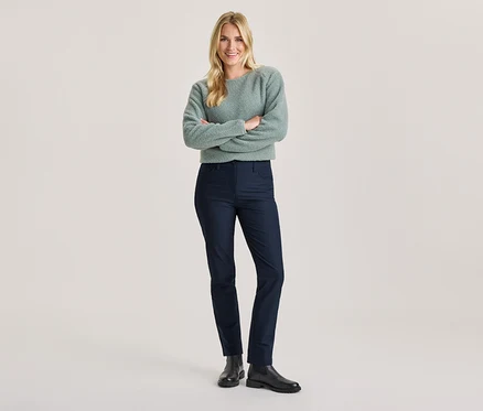 Femme souriante aux cheveux blonds, bras croisés, portant un pull duveteux vert clair, un pantalon bleu foncé et des bottes noires.