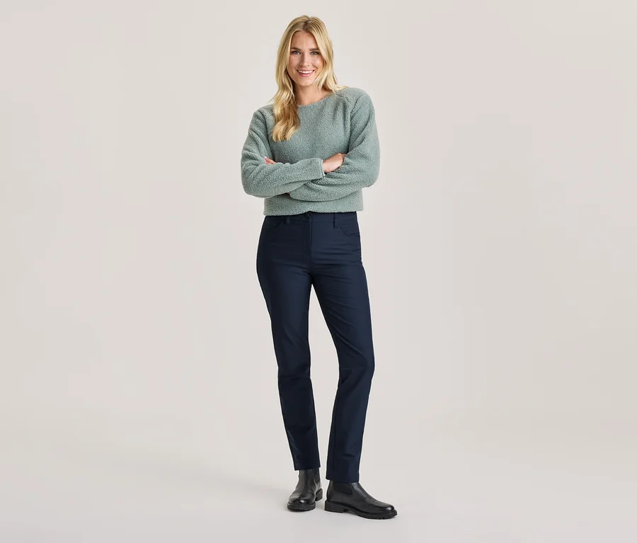 Femme souriante aux cheveux blonds, bras croisés, portant un pull duveteux vert clair, un pantalon bleu foncé et des bottes noires.