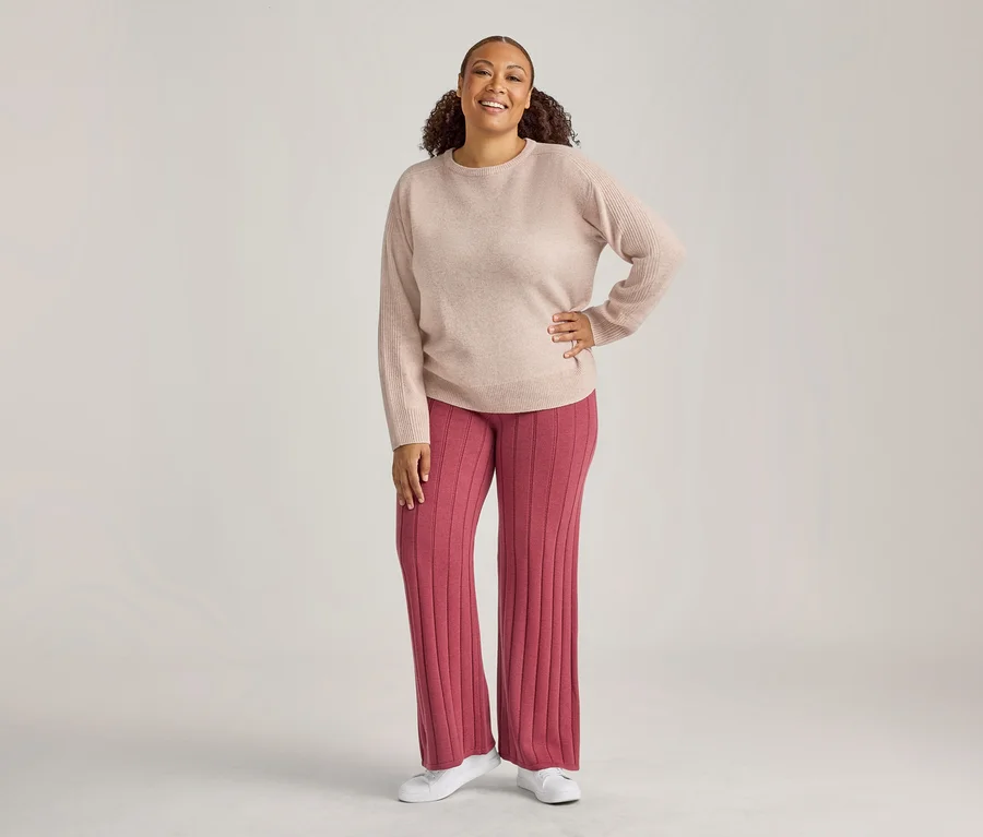 Femme souriante debout, portant un pull à manches longues beige clair, un pantalon large côtelé rose foncé et des chaussures blanches, une main sur la hanche.