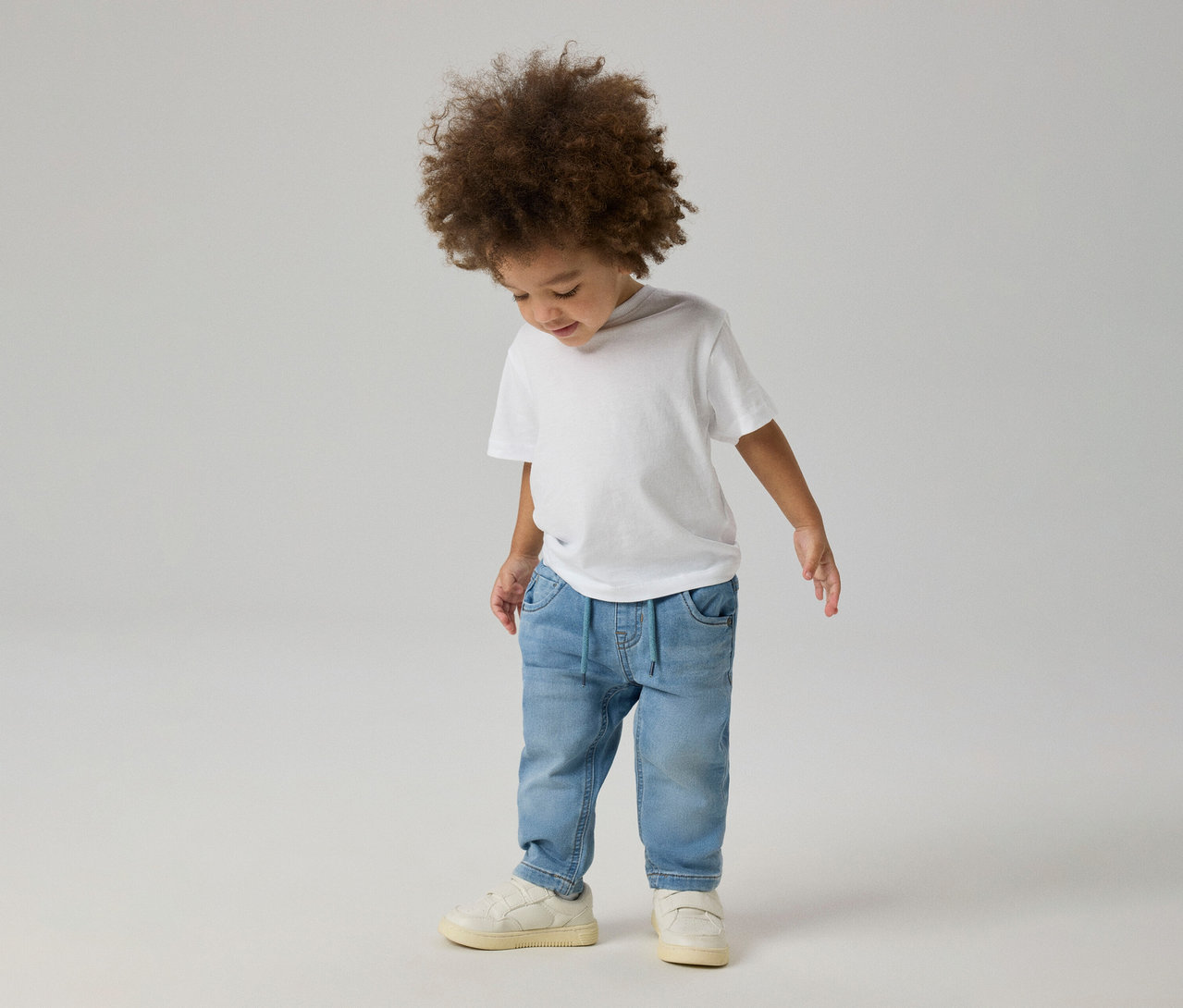 Jeune enfant aux cheveux bouclés portant un t-shirt blanc, un jean NAME IT Slim Sweat bleu clair et des baskets claires, regardant vers le bas.