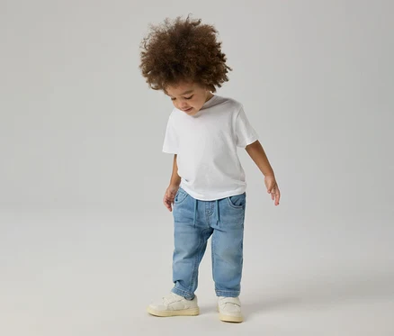 Jeune enfant aux cheveux bouclés portant un t-shirt blanc, un jean NAME IT Slim Sweat bleu clair et des baskets claires, regardant vers le bas.