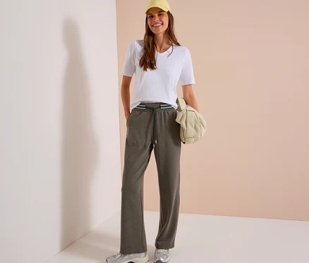 Femme souriante portant une casquette jaune, un t-shirt blanc et un pantalon en tissu CECIL gris foncé avec une taille rayée. Elle porte des baskets argentées et tient un sac rembourré crème.