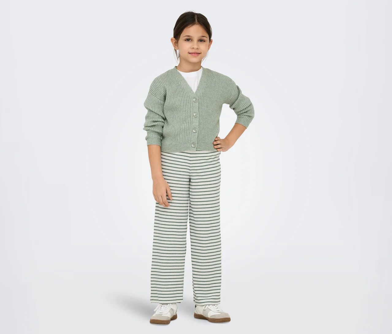 Jeune fille debout, les mains sur les hanches, portant un cardigan côtelé vert clair sur un t-shirt blanc, un pantalon large rayé et des baskets claires.