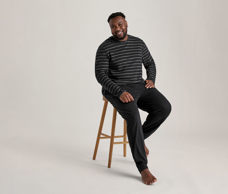 Homme souriant assis pieds nus sur un tabouret en bois, portant un ensemble pyjama rayé noir et gris.