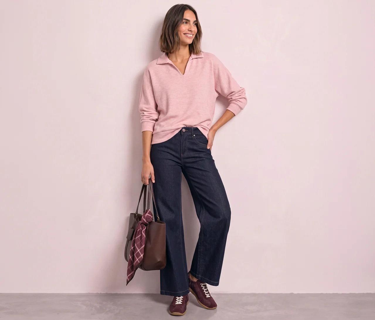 Une femme souriante aux cheveux bruns courts se tient debout, regardant vers la droite, vêtue d'un polo Street One rose clair, d'un jean large bleu foncé et de baskets bordeaux. Elle tient un sac cabas marron avec une écharpe à carreaux.