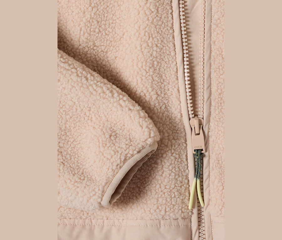 Gros plan d'une veste teddy CECIL beige, montrant la texture moelleuse, une fermeture éclair avec une tirette beige et un cordon vert et jaune, à côté d'un poignet côtelé.