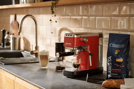Une machine à porte-filtre rouge « Lapressa » prépare du café dans un verre. À côté se trouvent un moulin à café et un paquet de café.