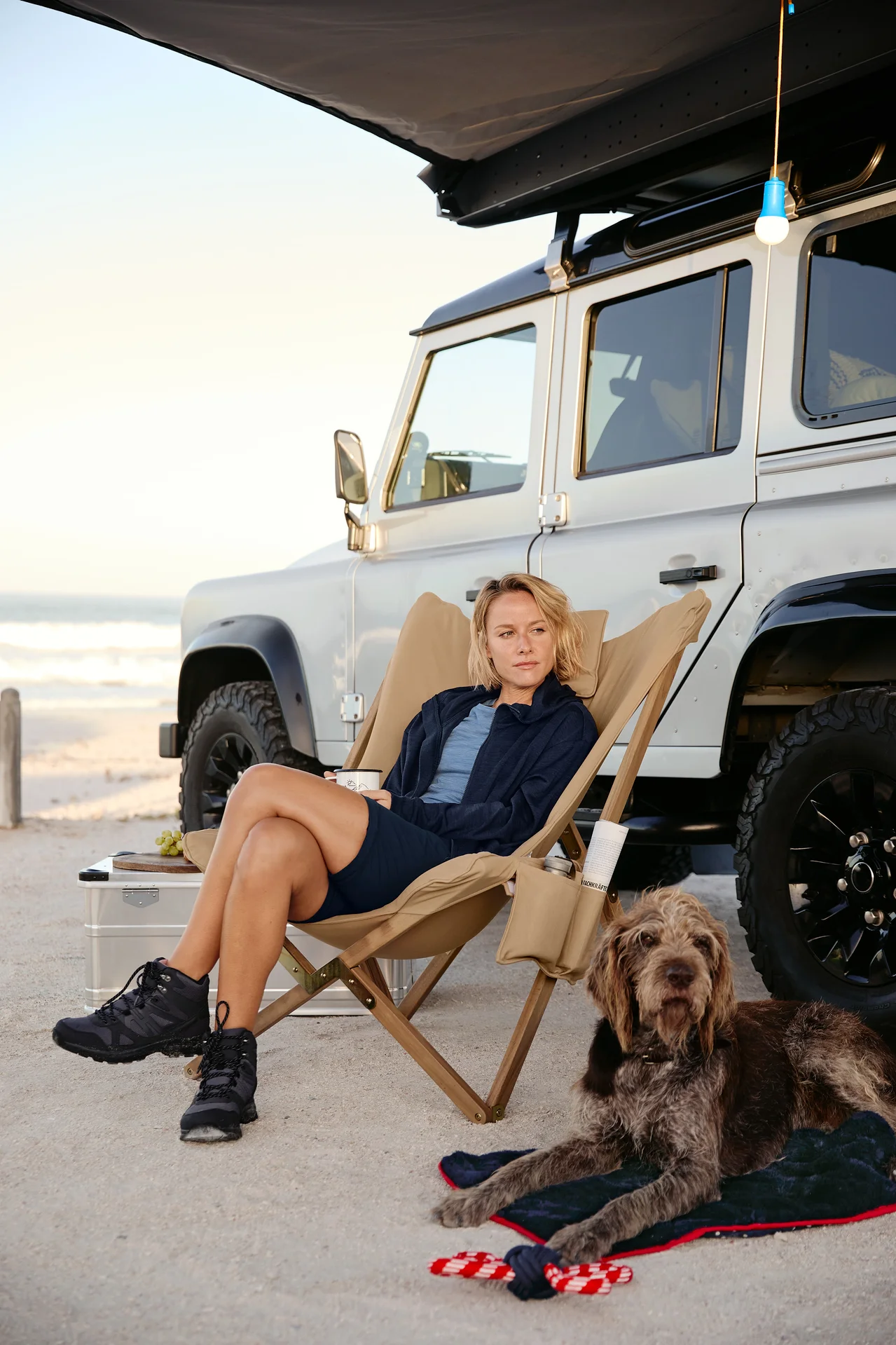 Une femme assise dans un fauteuil relax avec un mug, un chien est allongé à côté sur une couverture moelleuse pour chien avec un jouet. Une lampe de camping à LED, un pantalon fonctionnel 2 en 1 et une veste à capuche sont à l'arrière.