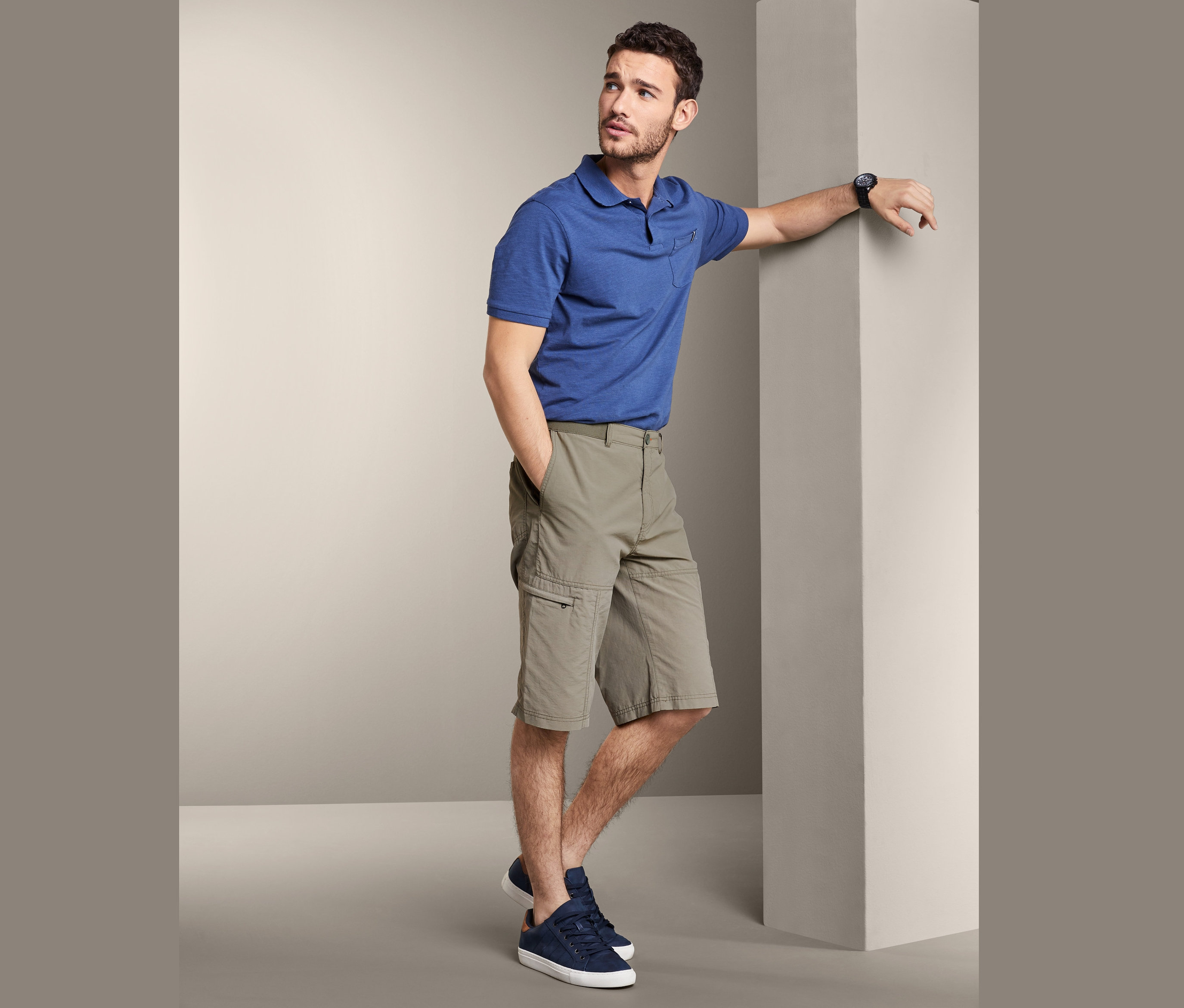 Un homme appuyé contre un mur porte un polo bleu et un bermuda.