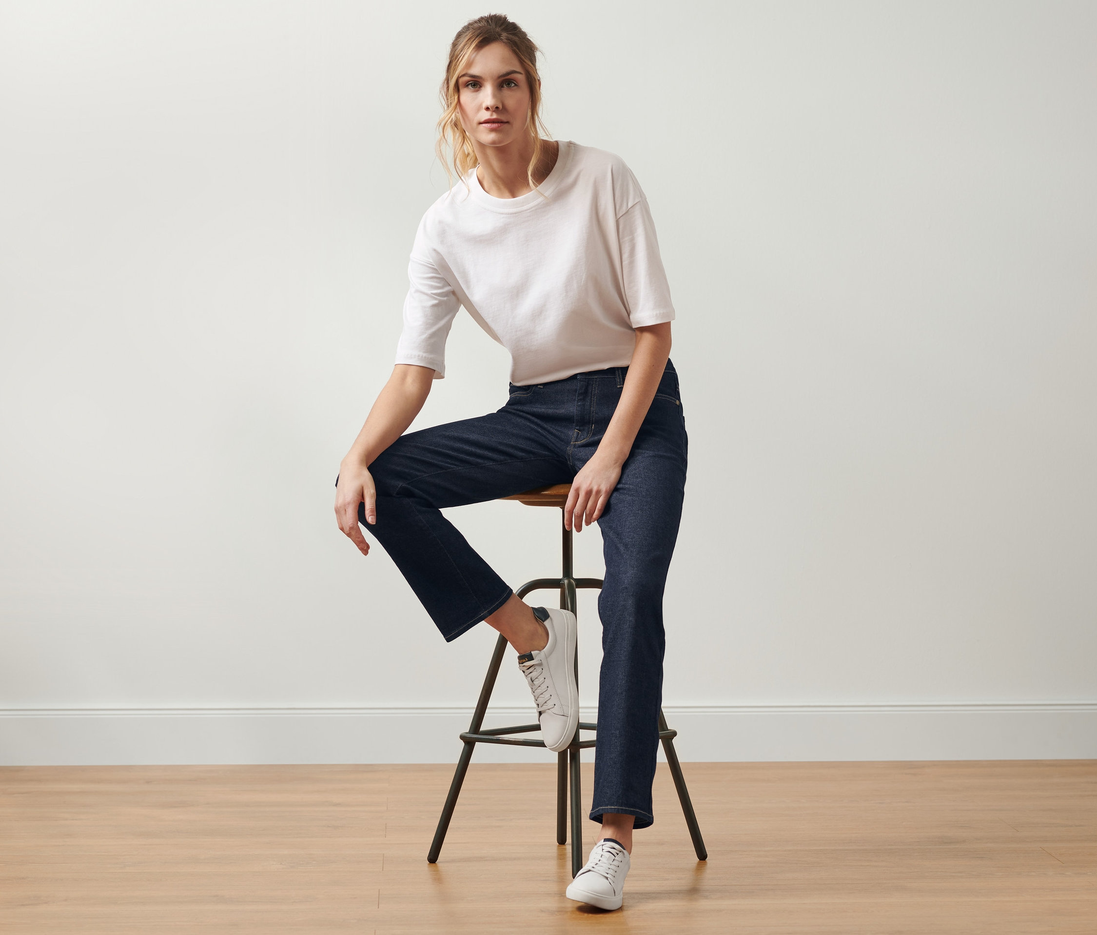 Une femme est assise sur un tabouret et porte un jean droit « Fit Kira », denim bleu foncé.