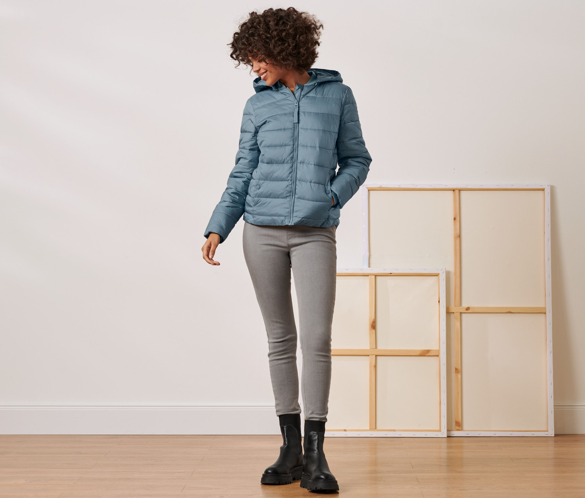 Femme posant en veste matelassée à capuche, jean gris et bottes noires.