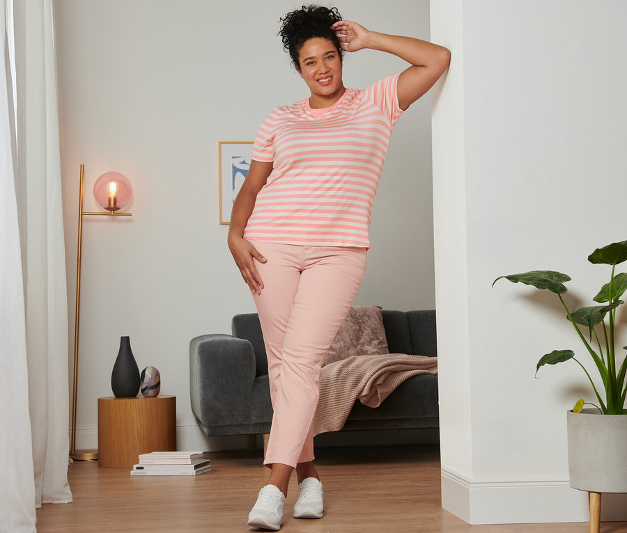 Une femme pose, appuyée contre un mur, portant un t-shirt à rayures rose, un jean slim rose et des baskets en cuir velours.