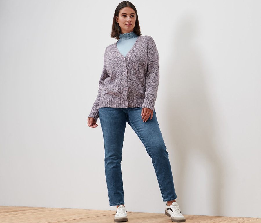 Femme posant en blouse à col montant bleu fumée, jean slim en denim « Fit Emma » bleu moyen, veste en maille avec de la laine et baskets en cuir.