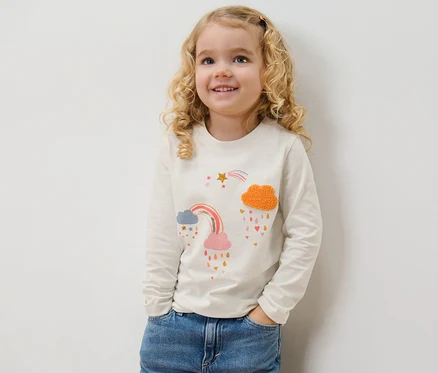 Une fillette souriante pose en jean et porte un t-shirt à manches longues avec un imprimé arc-en-ciel.