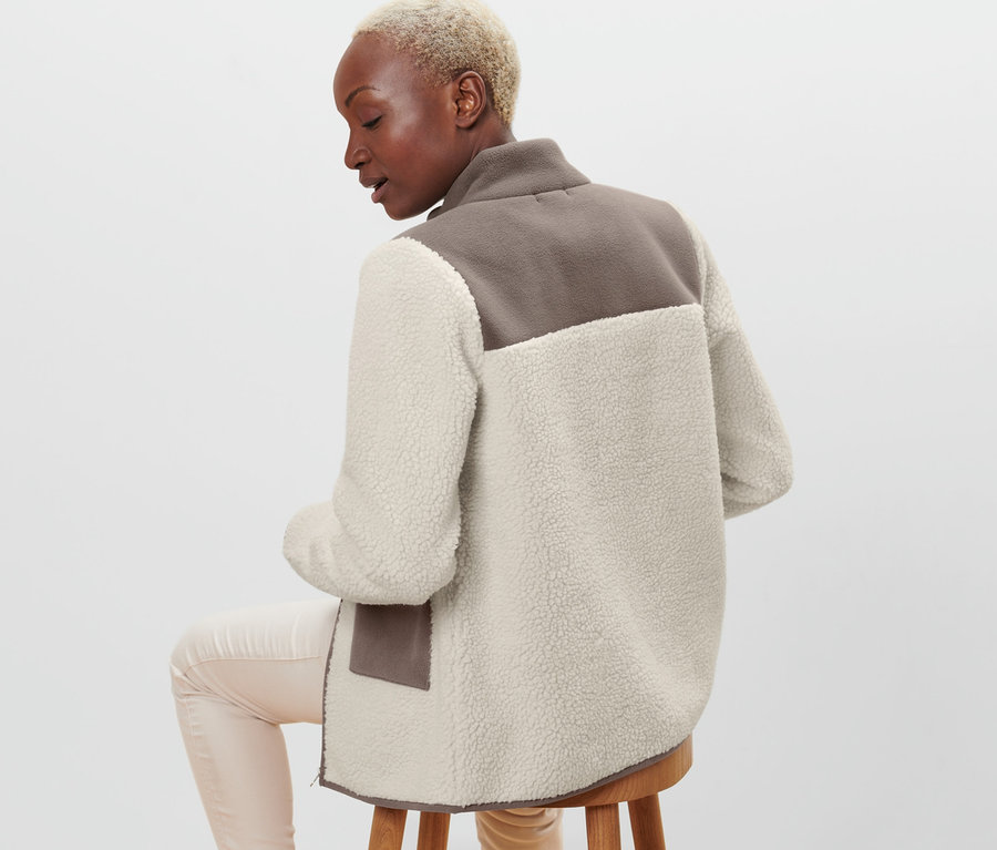 Une femme est assise sur un tabouret, le dos tourné à la caméra, et porte une veste en teddy beige et marron.