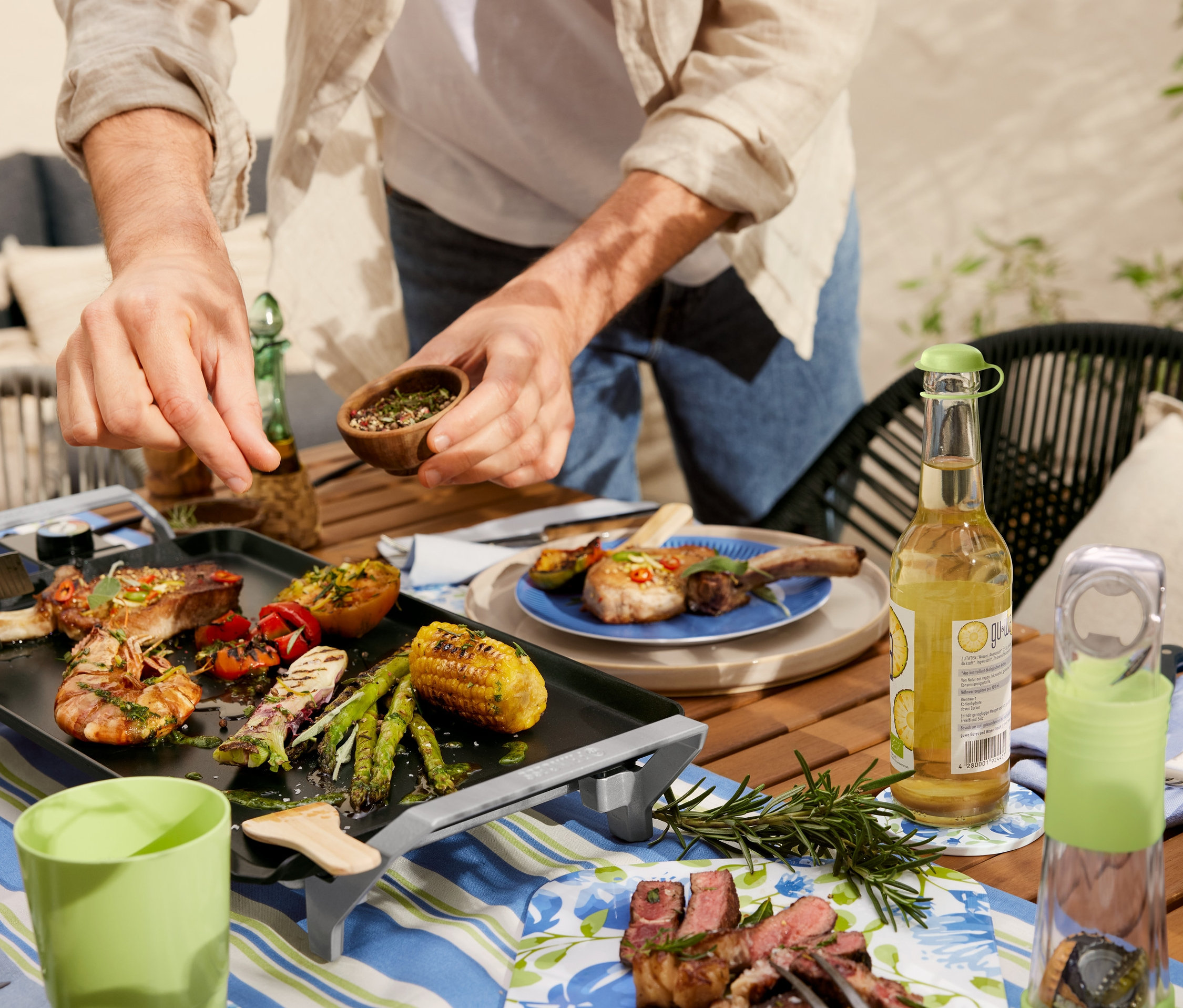 Un homme assaisonne des aliments sur un gril teppanyaki de table. Sur la table, il y a un chemin de table en coton rayé, un décapsuleur avec bac de récupération et une planche à servir.
