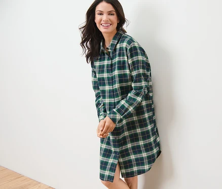 Une femme souriante, appuyée contre un mur blanc, porte une chemise de nuit en flanelle verte.