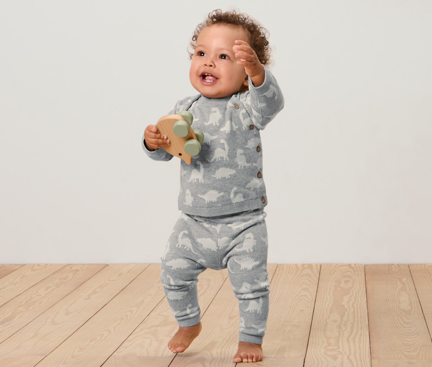 Un bébé se tient sur un plancher en bois et tient un jouet en bois. Il porte un ensemble en maille pour bébé.