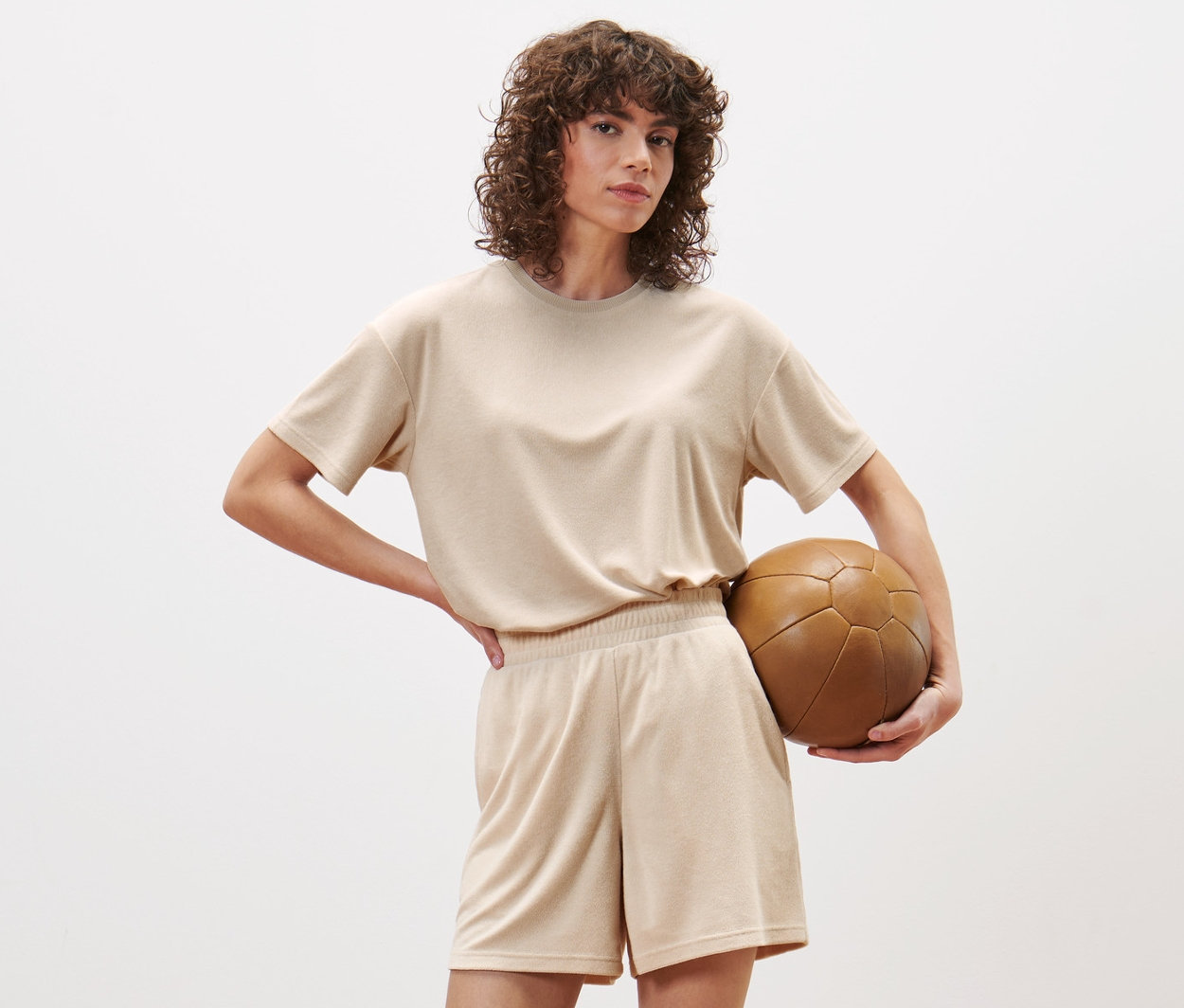 Une femme pose en studio, portant un short de sport en tissu éponge et tenant un ballon de foot marron.