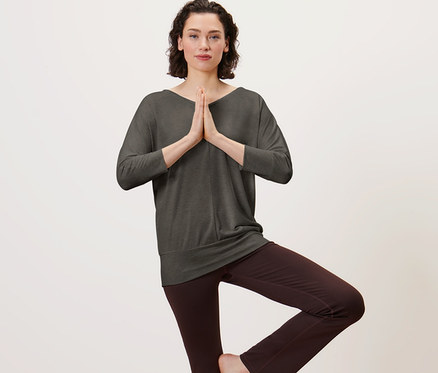 Femme portant un t-shirt de yoga olive debout sur un tapis de yoga en posture d'arbre.