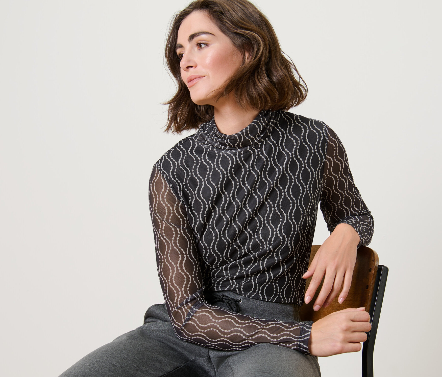 Une femme est assise sur une chaise et porte un haut manches longues à col roulé en mesh imprimé et un pantalon de survêtement gris.