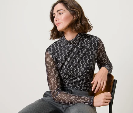 Une femme est assise sur une chaise et porte un haut manches longues à col roulé en mesh imprimé et un pantalon de survêtement gris.
