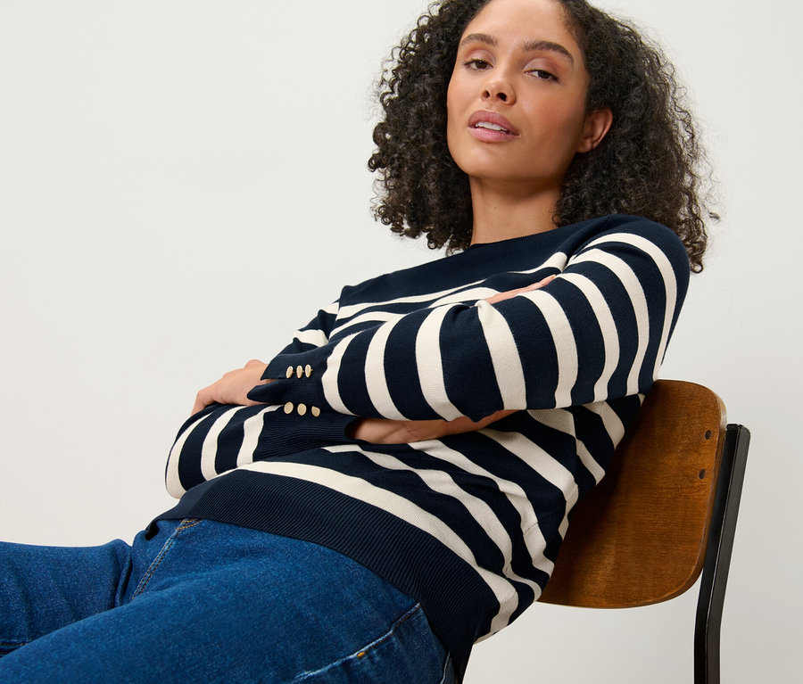 Une femme assise sur une chaise, les bras croisés, portant un pull en maille fine rayé bleu foncé.